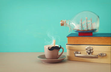 Image of cup of coffee, old book, old suitcase box and boat in the bottle over wooden table. Father's day concept.