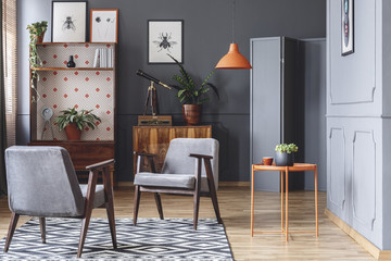 Two armchairs, orange coffee table, plants, wooden cupboards and grey walls in a living room...