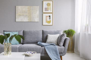 Real photo of a grey sofa standing in a stylish living room interior behind a white table with leaves and in front of a grey wall with posters - obrazy, fototapety, plakaty