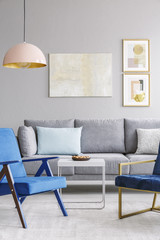 Real photo of two blue chairs standing in front of a grey couch in a elegant living room interior with posters in gold frames and hanging lamp