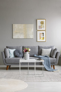 Real photo of a grey couch with cushions and blanket standing in living room interior in front of a wall with paintings and behind a white table