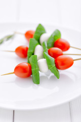 cherry tomatoes with basil and mozzarella cheese on .wooden skewers placed on a white plate and white wooden table