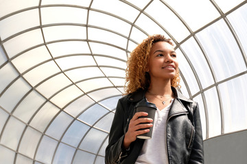 A young black woman on a walk in an urban environment. A modern beautiful African-American student girl in a leather jacket and white T-shirt is smiling and holding a glass in her hand. Copyspace.