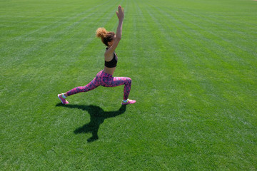 A young African-American girl in a black T-shirt, pink pants and sneakers doing sports exercises on green grass and raising her hands up. Modern style of life. Copy space.