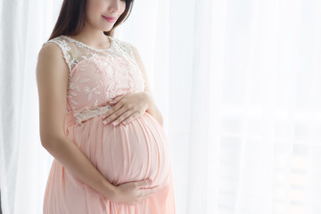 Pregnancy woman  in beautiful pink dress standing  close up a window in her bedroom.
