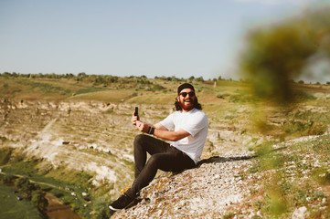 Smiling young bearded hipster man holding smartphone and smiling. Man sitting on rock