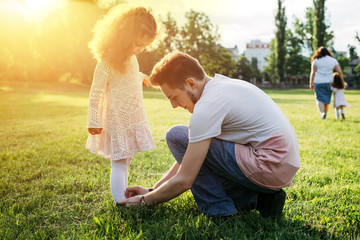 A father helping his little daughter with her shoes in the park. Cute curly kid girl in beautiful dress