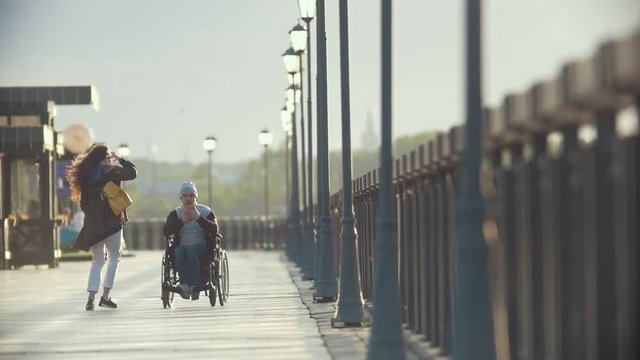 Disabled man in a wheelchair takes pictures of young woman at the quay