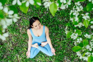 beautiful brunette woman in a sporty blue suit against a background of flowering spring trees sitting on green grass and doing yoga.View from above