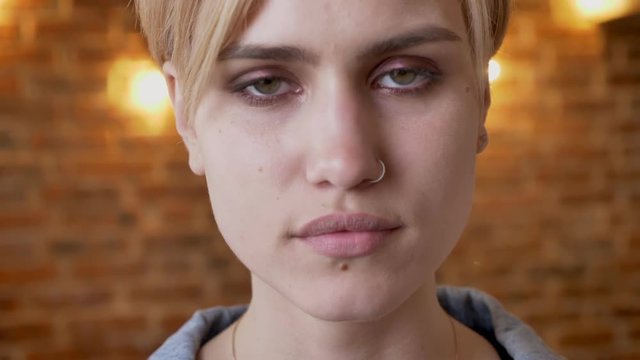 Young attractive serious hipster girl is watching at camera, brick background, closeup