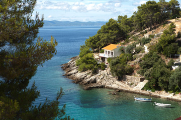 A picturesque view of a small house with a tiled roof on a rocky coast of turquoise sea with boats
