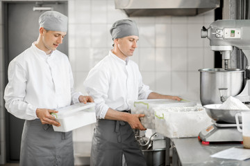 Two male confectioners holding containers with food ingredients standing at the bakery manufacturing