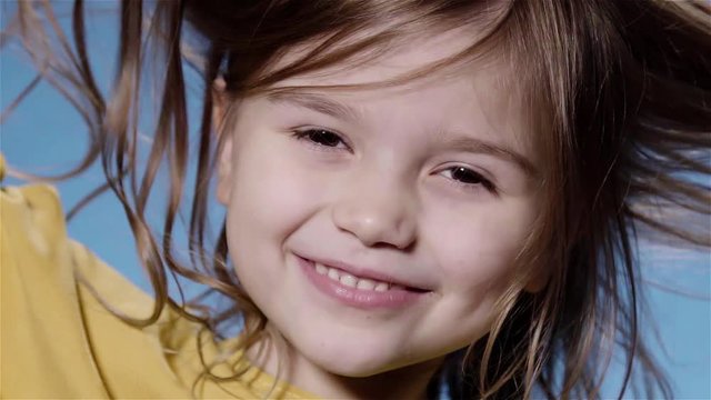 Portrait of a funny cute little girl with long dark fair hair wearing a yellow sweater playing with her hair and looking at camera. Blue background. Super slow motion locked down close up shot