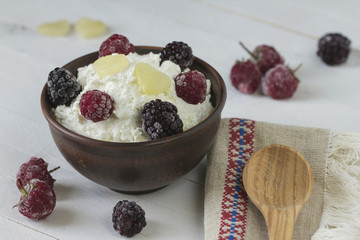 Cottage cheese in a brown ceramic bowl with frozen raspberries and blackberries with authentic flax embroidered napkin and a wooden spoon on a white wooden background.  Close-up.