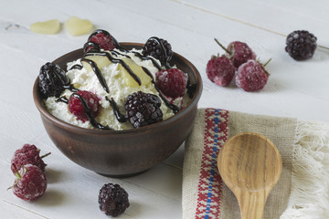 Cottage cheese in a brown ceramic bowl with frozen raspberries and blackberries with authentic flax embroidered napkin and a wooden spoon on a white wooden background.  Close-up.