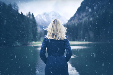 Woman in blue coat standing at lake during snowfall. Selective focus used.