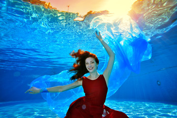 Obraz na płótnie Canvas The model girl with red hair sits and poses underwater at the bottom of the pool in a red dress on a blue background. She looks at the camera and smiles. Portrait. Landscape image orientation