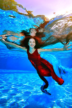 A girl with red hair swims and poses underwater in the pool on a blue background. She spread her hands to the sides and smiles. Portrait. Vertical orientation of the image