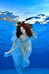 A beautiful bride with red hair is swimming in a white transparent dress under the water in the pool on a blue background. Portrait. Vertical orientation