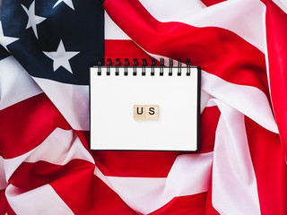 Notepad with blank page, wooden letters of the alphabet in the form of the word US on the background of the US Flag. Top view, close-up. The concept of an independent, strong country and nation