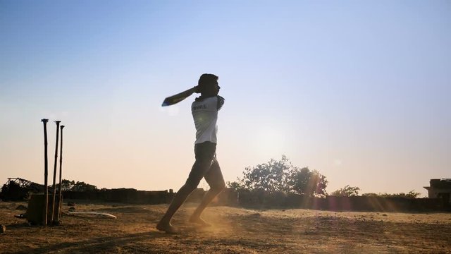 A young boy batting hits the cricket ball for a six in a open field of Indian village. A slow motion shot of a young teenager playing cricket against the sun strikes the cricket ball hard