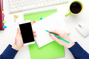 white desk, male hands, man uses white smart phone, guy writes in a Notepad, office supplies,  white background with copy space, for advertisement, top view,