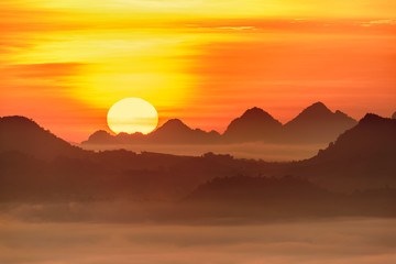 Fototapeta na wymiar Distant scenic view at dawn / scene of evanescent atmosphere in the evergreen forest. Big sun rises behind a mountain range in the morning. Dense morning fog or mist rolling in over lush wilderness.