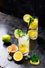 Lemon fruit lime slices caipirinha from Brazil, lemonade mint ice cubes in cold glasses on dark background, alcoholic mojito cockail ice tea green mint leaves, brown sugar spoon, copy space