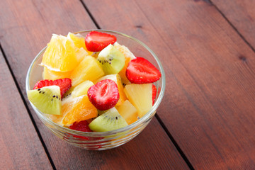 Fruit salad from pineapple, kiwi, orange, strawberry. Multicolored sliced fruit in a transparent dish. Vegetarian food on a wooden background. Citrus fruits for Asian cuisine