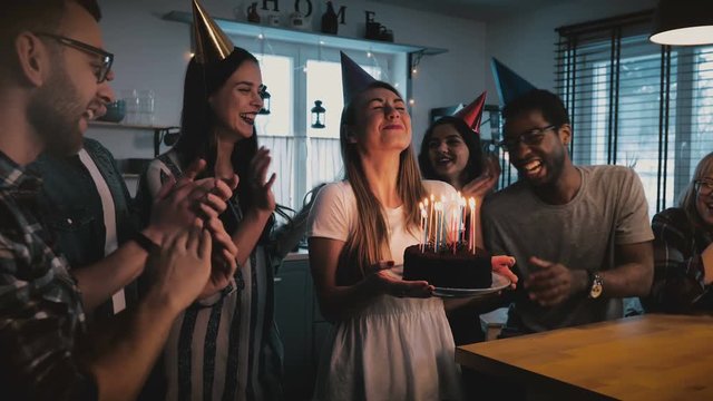 Happy European girl holding birthday cake, making a wish and blowing, multiethnic friends sing at fun party slow motion.