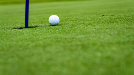 Side view of a golf ball on a sloped putting green, close to the hole with a blue pin
