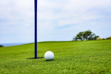 Obraz premium Side view of a golf ball on a sloped putting green, close to the hole with a blue pin, with sky and clouds, and a tree in the background 