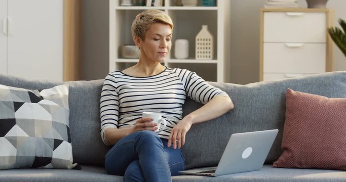 Caucasian middle aged woman drinking tea or coffee from a white cup while watching something on the laptop computer on the couch in the living room. Inside