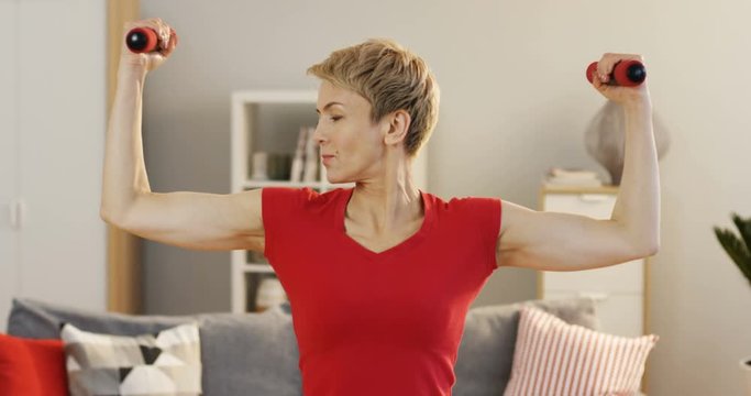 Caucasian middle aged woman swinging her hands with dumbbells at home. Indoors