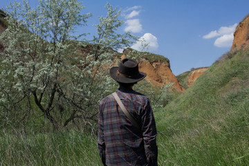 Back view of young male hiker in cowboy hat walking outdoors in summer day. Hipster traveler.
