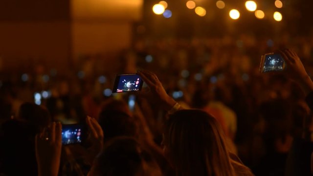 People are taking pictures of musical concert with phones, standing in dark room. Crowd captures exciting event, using smartphones, holding gadgets in hands in front of stage. Men, women have good