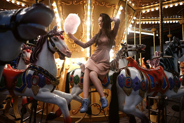 Young beauty model woman posing with old horse carousel in summer park with magic lights