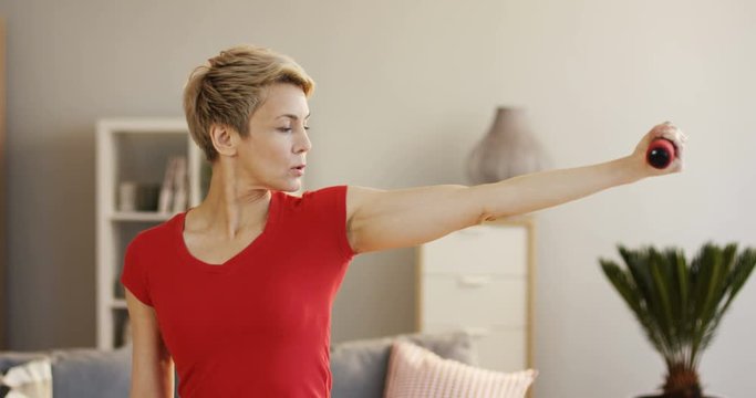 Portrait shot of the Caucasian middle aged woman with short blond hair exercising with dumbbells in front of the camera at home. Indoor