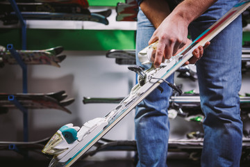 Theme of business sale and service of ski equipment. The hands of young Caucasian man have mountain skis, checks and adjusts the fastening for boot. On the wall of the stand with skis in the store