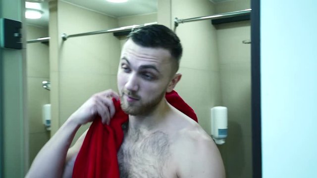 Portrait of young man, who is going out from the shower in sport center. Handsome sportsman with stylish haircut and beard is moving to his locker to dress up and drying his head by the red towel on