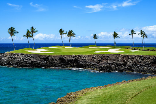 Sunny day on a tropical golf course fairway with the putting green in the distance surrounded by palm trees and sand traps, lava rock, blue pacific ocean, and blue sky and white clouds in background