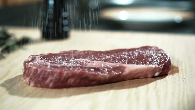 Close up of delicious steak is being prepared before cooking in luxury restaurant, slow motion. Piece of red meat is lying on wooden cutting board near green hearbs and being strewed with salt and