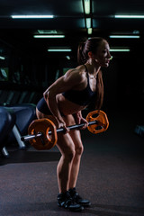 A strong, muscular, girl in black shorts, performs an exercise of deadlift, with a sports bar. Dark gym.