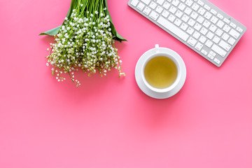 Cosy spring work desk concept. Computer keyboard, cup of tea and bouquet of lily of the valley flowers on pastel pink background top view copy space