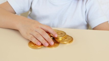 The child's hands are playing with gold coins of bitcoins on the table. The child plays with a crypto currency. Close-up.