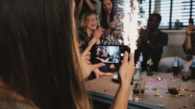 Girl taking a smartphone photo of birthday party with sparkling firework and multiethnic friends clapping slow motion.