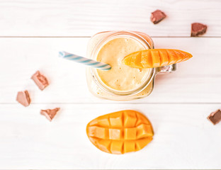 summer drink mango milkshake in mason jar with drinking straw decorated with a half of mango, chocolate pieces flat lay top view on white rustic background