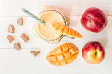 summer drink mango milkshake in mason jar with drinking straw decorated with a half of mango, chocolate pieces and nectarines flat lay top view on white rustic background