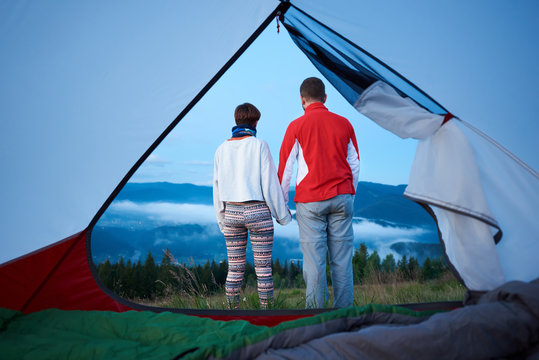 Rear view of two young people holding hands enjoy the morning scenery of the mountains on which the fog descended. View from inside a tent