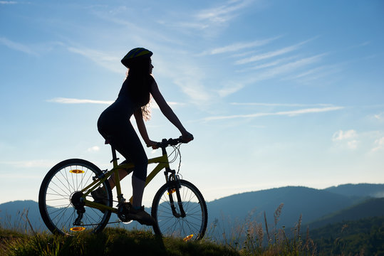 Silhouette of strong female biker cycling on mountain bike in the sunny morning, wearing helmet, against blue sky. Outdoor sport activity, lifestyle concept. Copy space